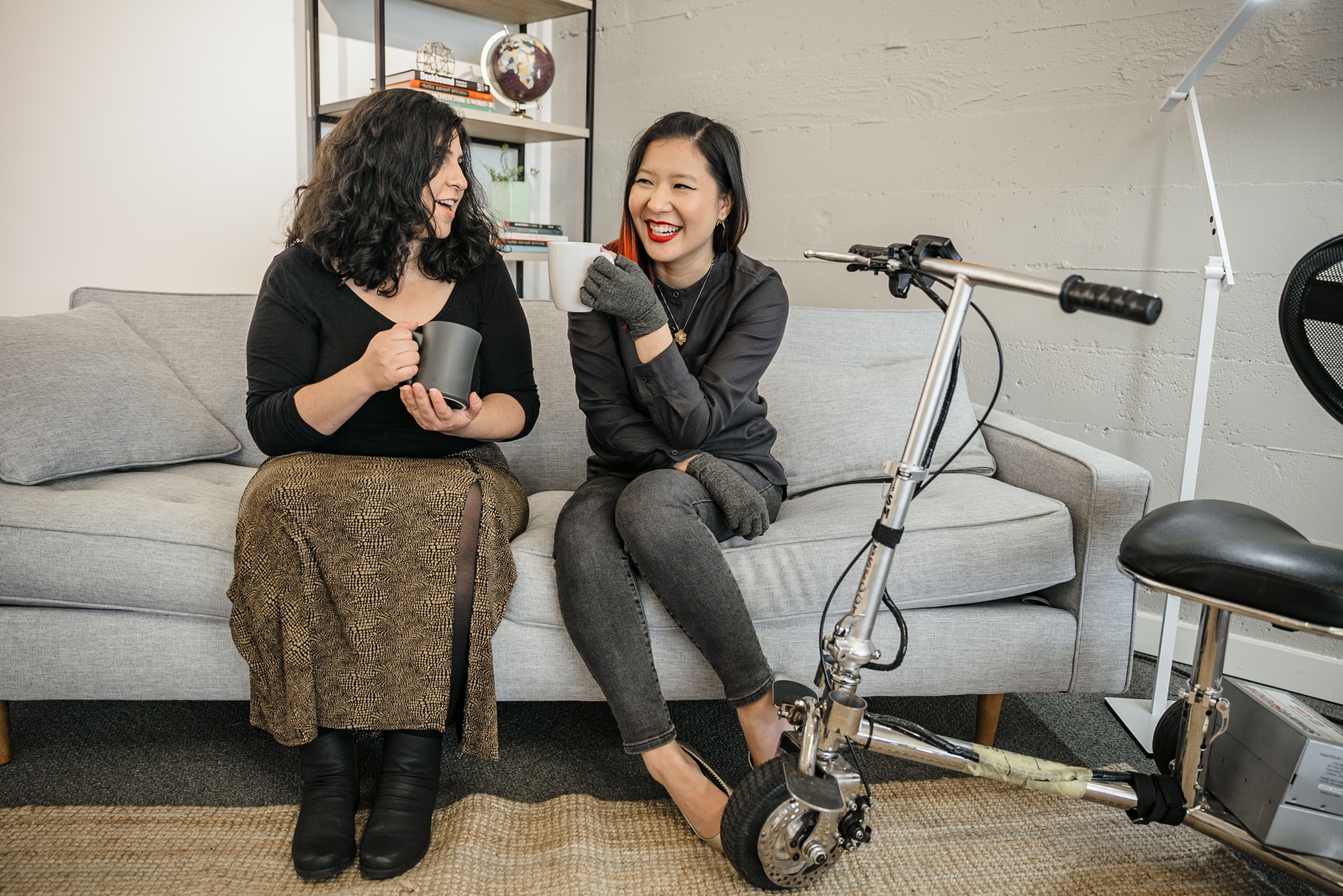 A Latinx disabled woman and an Asian disabled genderfluid person chat with coffee on a couch. An electric mobility scooter rests on the side.